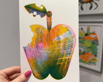May include: A colorful print of a bitten apple with a leaf. The apple is a mix of yellow, orange, pink, and green, with a bite taken out of the side. The leaf is green and blue. The artwork is on white paper.