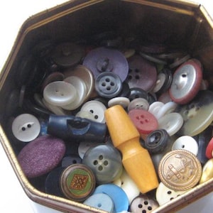 May include: A hexagonal metal tin filled with a variety of loose buttons in assorted shapes, sizes, and colours, including white, black, red, purple, blue, and metallic tones, featuring a prominent light brown wooden toggle button on top.