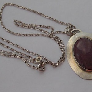 May include: A silver-toned metal necklace featuring a long, oval-shaped pendant with a smooth, dark maroon-coloured stone set in the centre. The pendant is attached to a delicate, thin chain with a circular clasp mechanism visible near the pendant.