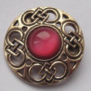 May include: A circular brooch in a gold-tone metal. It has a Celtic knot design around a central, red gemstone. This vintage brooch is a decorative accessory, suitable for adding a touch of history to an outfit.