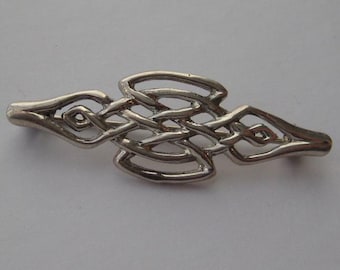 May include: A silver-toned Celtic knot barrette. The barrette features an intricate, openwork design of interwoven lines and shapes. The barrette is symmetrical, with a central knot and tapering ends. The metal has a polished finish, reflecting light.
