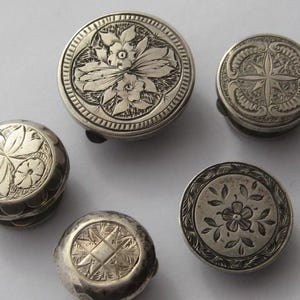 May include: A selection of antique silver pill boxes, each with a unique engraved design. The round boxes feature hinged lids and are adorned with floral and geometric patterns. The silver has a slightly aged appearance, with detailed engravings of flowers and starbursts.