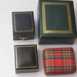 May include: Four jewellery boxes of varying sizes and designs. One large dark green box with gold trim, one small black box with gold trim, one small dark blue box with gold trim and a metal latch, and one rectangular box with a red plaid pattern.