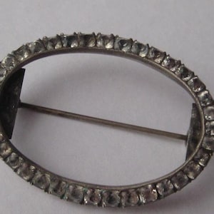 Op de afbeelding: Ovale zilveren broche met een rij heldere, vierkant geslepen stenen langs de rand. De broche heeft een speld aan de achterkant om vast te maken. Het ontwerp is eenvoudig en elegant, geschikt voor diverse gelegenheden. Ongeveer 3 cm lang.