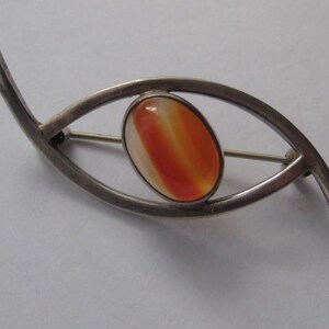 May include: Silver brooch featuring an orange and white agate stone in the shape of an eye.