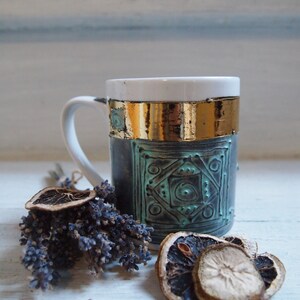 Puede incluir: Una taza de cer&aacute;mica blanca con un dise&ntilde;o geom&eacute;trico negro y turquesa y detalles dorados. La taza est&aacute; sobre una superficie blanca con lavanda seca y rodajas de naranja.