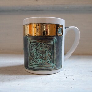Puede incluir: Una taza de cer&aacute;mica blanca con una banda dorada y un dise&ntilde;o geom&eacute;trico azul verdoso. La taza tiene una asa redondeada.