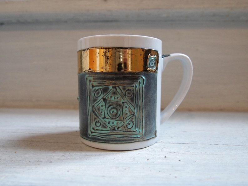 Puede incluir: Una taza de cer&aacute;mica blanca con una banda dorada y un dise&ntilde;o geom&eacute;trico azul verdoso. La taza tiene una asa redondeada.