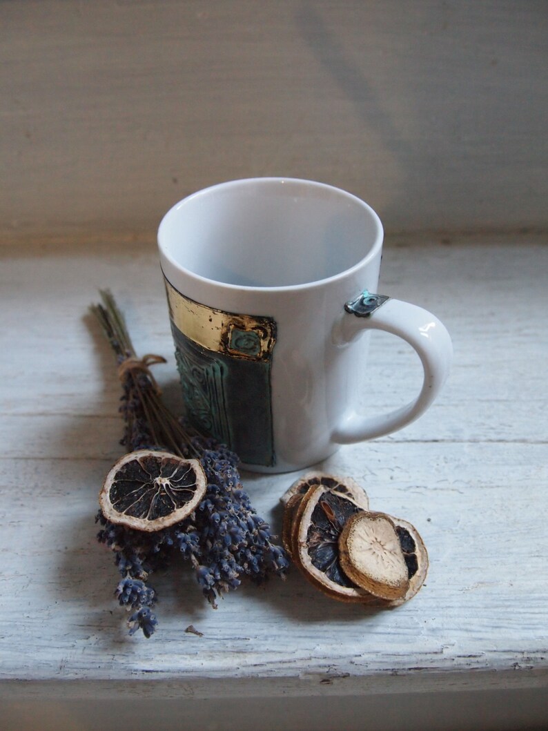 Puede incluir: Taza de cer&aacute;mica blanca con un dise&ntilde;o azul y dorado. La taza est&aacute; sobre una superficie de madera blanca con lavanda seca y rodajas de naranja.