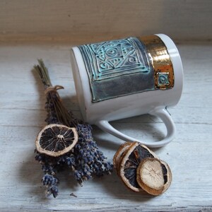 Puede incluir: Una taza de cer&aacute;mica blanca con un dise&ntilde;o geom&eacute;trico azul y dorado. La taza tiene un asa y est&aacute; sentada sobre una superficie de madera blanca con lavanda seca y rodajas de lim&oacute;n.