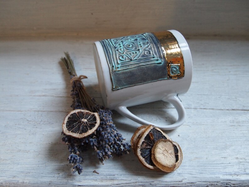 Puede incluir: Una taza de cer&aacute;mica blanca con un dise&ntilde;o geom&eacute;trico azul y dorado. La taza tiene un asa y est&aacute; sentada sobre una superficie de madera blanca con lavanda seca y rodajas de lim&oacute;n.