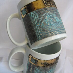 Puede incluir: Dos tazas de cer&aacute;mica blanca con dise&ntilde;os dorados y turquesa. La taza de arriba tiene el texto "Konstanz University" en la banda dorada. La taza de abajo tiene el texto "The University" en la banda dorada.