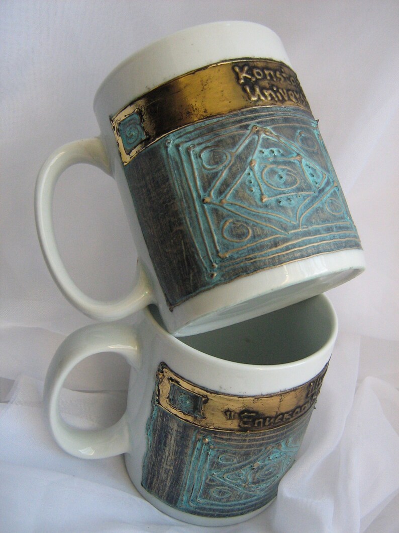 Puede incluir: Dos tazas de cer&aacute;mica blanca con dise&ntilde;os dorados y turquesa. La taza de arriba tiene el texto "Konstanz University" en la banda dorada. La taza de abajo tiene el texto "The University" en la banda dorada.