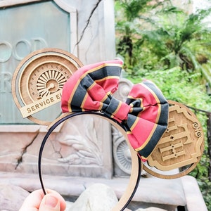 May include: A pair of brown and black mouse ears with a red and black striped bow. The ears feature a wooden design with the text "The Hollywood Tower Hotel" and a circular design with the text "Service Elevator".