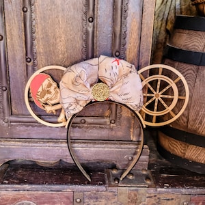 May include: A pair of brown and gold Mickey Mouse ears with a pirate skull and treasure map design. The ears are on a wooden background.
