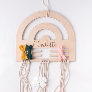 Puede incluir: Un soporte para lazos para el pelo de madera con forma de arcoíris con el nombre "Charlotte" grabado en él. El soporte tiene múltiples cuerdas que cuelgan de él, y hay varios lazos para el pelo de colores que cuelgan de las cuerdas.