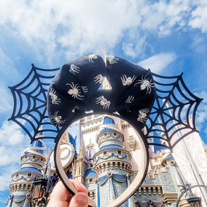 Könnte beinhalten: Ein schwarz-weißes Spinnennetz-Haarband mit einer schwarzen Schleife und weißen Spinnen. Das Haarband wird vor dem Schloss von Aschenputtel in Walt Disney World gehalten.