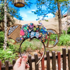 May include: A pair of wooden Mickey Mouse ears with a colourful bow. The left ear features a wooden cart with a pile of gold coins. The right ear features a wooden compass with an anchor.