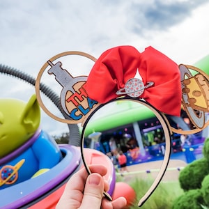 May include: A pair of light brown mouse ears with a red bow and a silver charm. The left ear features a clear plastic cutout of a beaker with the text "The Claw" inside. The right ear features a clear plastic cutout of a pizza slice with a silver ring around it.