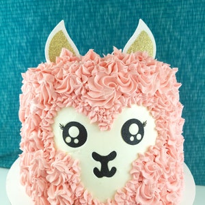 May include: A square cake decorated to resemble a llama. The cake features pink frosting, a white face with black eyes and a mouth, and gold glitter ears. The cake is on a white cake stand.