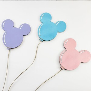 K&ouml;nnte beinhalten: Drei Mickey-Maus-f&ouml;rmige Ballons aus Holz, einer hellblau, einer helllila und einer hellrosa, mit Schn&uuml;ren befestigt.