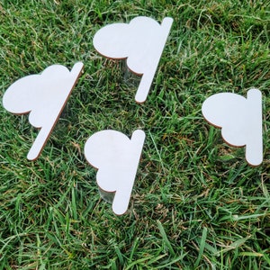 May include: Four white wooden cloud-shaped ornaments with a smooth, unfinished surface. The ornaments are arranged on a bed of green grass.