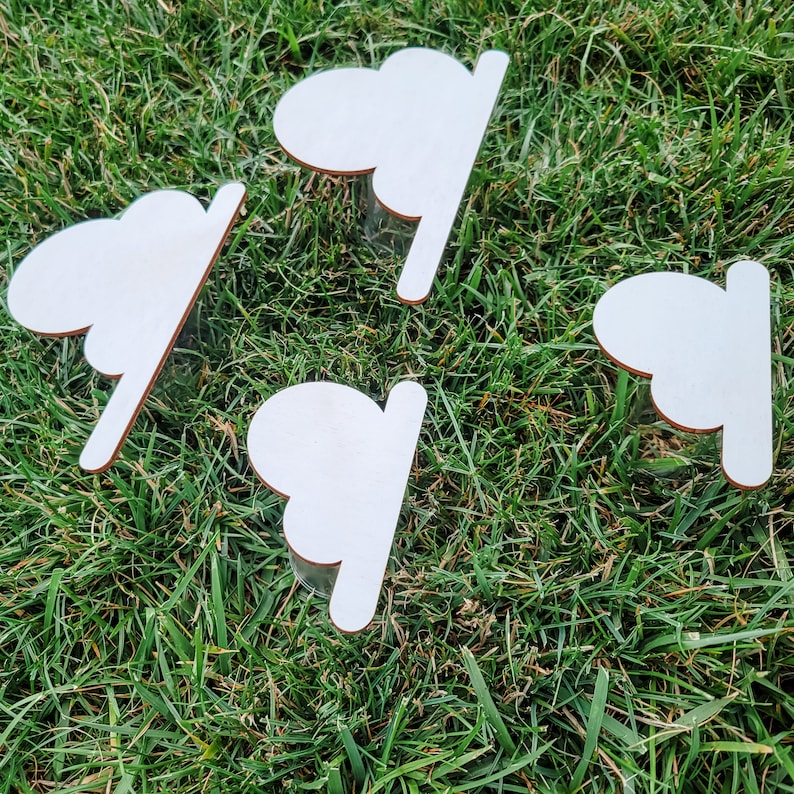 May include: Four white wooden cloud-shaped ornaments with a smooth, unfinished surface. The ornaments are arranged on a bed of green grass.