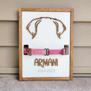 May include: A wooden plaque with a cutout of a dog's head, a pink dog collar, and the name "ARMANI" and the dates "2014-2023" written on it.
