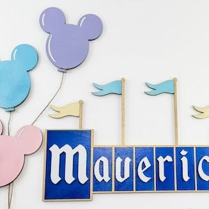 May include: Wooden name sign with the name "Maverick" in white letters on a blue background. The sign is decorated with three flags and three Mickey Mouse-shaped balloons in pink, blue, and purple.