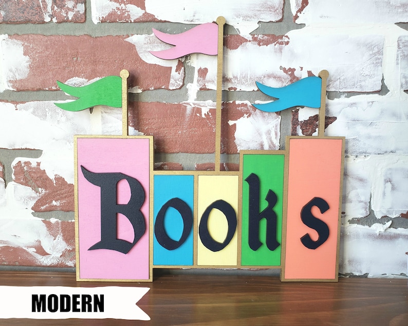 May include: A colorful wooden bookend with the word "Books" in black gothic font. The bookend features pink, blue, yellow, green, and orange blocks, with small flags on top. The word "MODERN" is printed at the bottom.