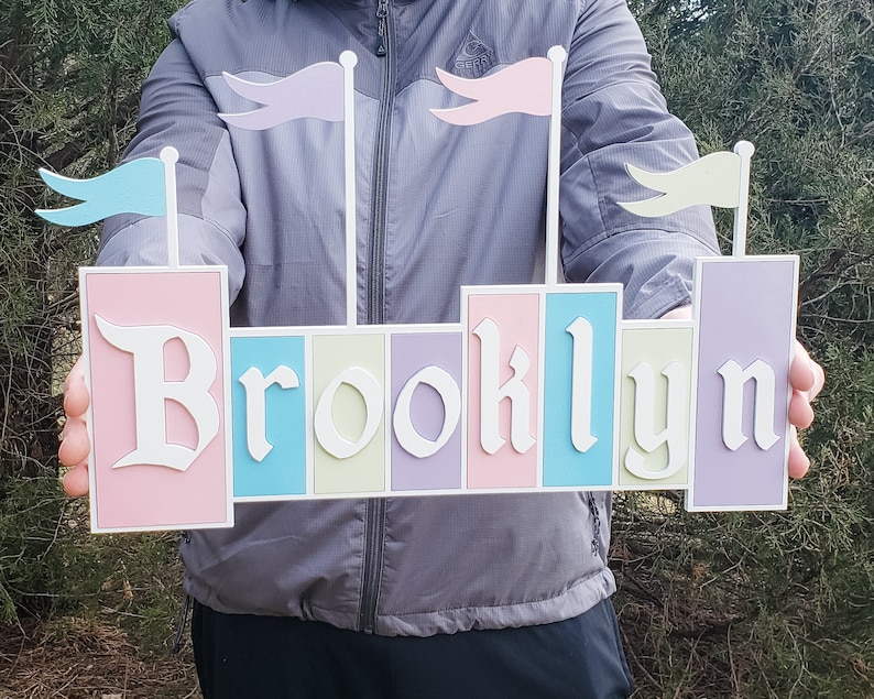 May include: A pink, blue, green, and purple wooden sign with the name "Brooklyn" spelled out in white letters. The sign has flags on top of each letter.