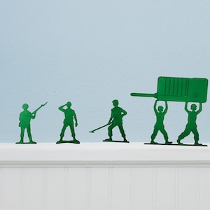 Puede incluir: Soldados de juguete de pl&aacute;stico verde en varias poses, incluido uno apuntando con un rifle y dos levantando un gran walkie-talkie verde. Los juguetes est&aacute;n colocados en un estante blanco sobre un fondo azul claro.