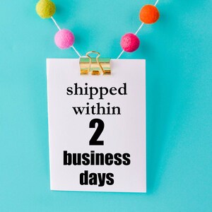 May include: A white card with a gold paper clip hanging from a string of colorful pom-poms against a blue background. The card says "shipped within 2 business days".