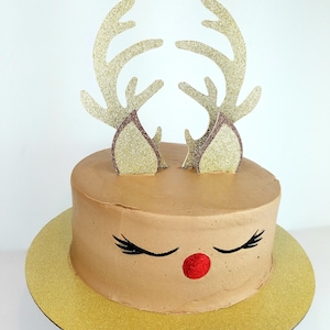 May include: A chocolate cake decorated with a reindeer face, including antlers, eyes, and a red nose. The cake is on a gold cake board.