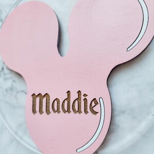 May include: A pink wooden Mickey Mouse head-shaped decoration with the name "Maddie" engraved in a brown font. The design includes white accents and is set against a blurred background.
