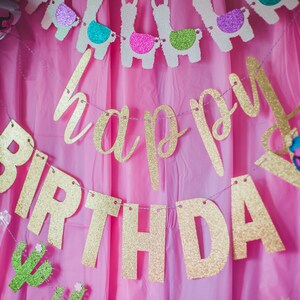 May include: A pink backdrop with a gold glitter "Happy Birthday" banner and llama-themed decorations.