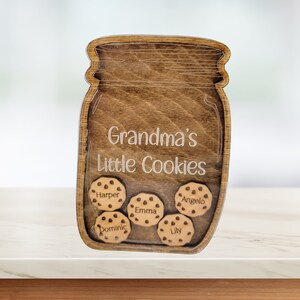 May include: Wooden jar-shaped sign with the text "Grandma's Little Cookies." Inside the jar are five wooden cookies, each with a different name: Harper, Dominic, Emma, Lily, and Angelo. The sign is made of stained wood.