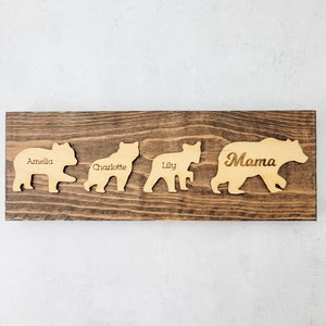Könnte beinhalten: Ein Holzschild mit fünf Bärensilhouetten, jede mit einem eingravierten Namen. Die Namen sind Amelia, Charlotte, Lily, Mama. Das Schild ist braun und hat einen rustikalen Look.