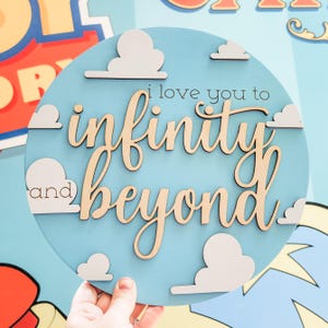 May include: A round, light blue wooden sign with the phrase "I love you to infinity and beyond" in a mix of fonts. White cloud cutouts are scattered around the text. The sign is held by a hand.