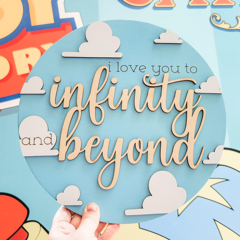May include: A round, light blue wooden sign with the phrase "I love you to infinity and beyond" in a mix of fonts. White cloud cutouts are scattered around the text. The sign is held by a hand.