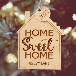 May include: A wooden house-shaped ornament with the words "HOME SWEET HOME" and "85 IVY LANE" engraved. A small key-shaped tag with "2021" is attached. The ornament is light brown and hangs against a blurred background of Christmas lights.