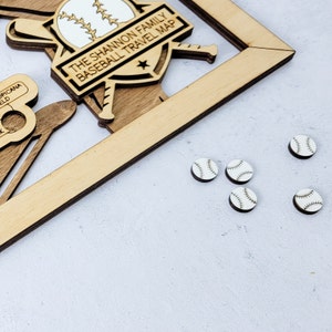 Puede incluir: Mapa de viaje con temática de béisbol de madera con el texto "THE SHANNON FAMILY BASEBALL TRAVEL MAP". El mapa incluye un diseño de bate y pelota de béisbol, junto con pequeños marcadores en forma de pelota de béisbol. El marco es de madera clara.