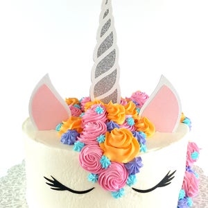 May include: A white cake decorated with pink, orange, and blue frosting swirls, topped with a silver unicorn horn and pink ears. The cake has black eyelashes painted on the frosting.