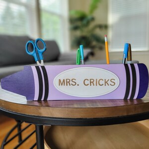 Könnte beinhalten: Lilafarbener Bleistifthalter in Form eines Buntstiftes mit schwarzen Streifen und einem weißen ovalen Etikett mit der Aufschrift "MRS. CRICKS".