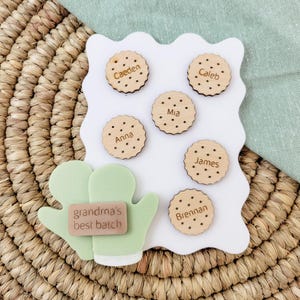 Puede incluir: Un conjunto de imanes personalizados de madera con forma de galleta con nombres: Caeden, Caleb, Mia, Anna, James y Brennan. Incluye un guante de cocina verde claro con el texto "grandma's best batch".