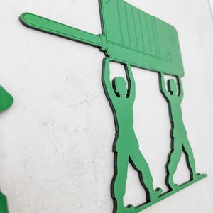 Puede incluir: Arte de pared de madera verde que representa dos siluetas de soldados de juguete levantando un gran rect&aacute;ngulo verde. Los soldados est&aacute;n en una pose cl&aacute;sica, sosteniendo la pieza rectangular sobre sus cabezas. El arte est&aacute; montado en una pared blanca.