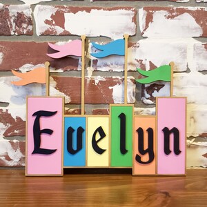 May include: A colorful wooden name sign spelling "Evelyn" in black gothic font. The sign features pink, yellow, green, and orange blocks with small flags on top. The background is a brick wall.