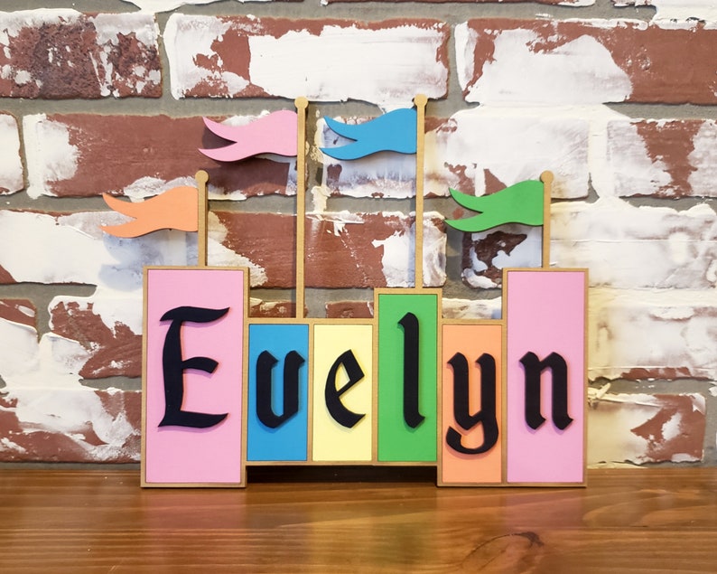 May include: A colorful wooden name sign spelling "Evelyn" in black gothic font. The sign features pink, yellow, green, and orange blocks with small flags on top. The background is a brick wall.