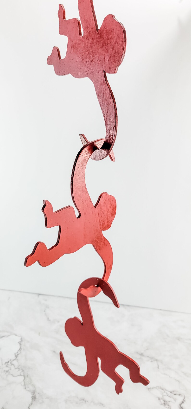 Puede incluir: Un m&oacute;vil de madera rojo con tres monos colgados unos de otros. Los monos est&aacute;n en una pose juguetona con los brazos y las piernas extendidos.