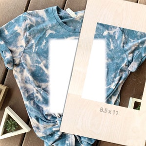 May include: A blue and white tie-dye t-shirt with a blank white rectangle in the center. The t-shirt is laid out on a wooden surface with a piece of wood with the text "8.5 x 11" printed on it.